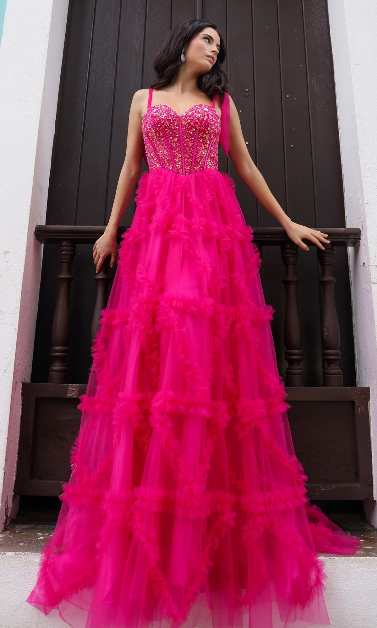 A woman stands before a dark wooden door wearing the Shoulder-Bows Long Ruffled Prom Gown T1340, a bright pink, floor-length tulle dress featuring floral details, ruffles, and a fitted beaded corset bodice.